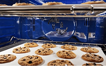 The blue interior of a wall oven, with two racks of chocolate chip cookies baking.