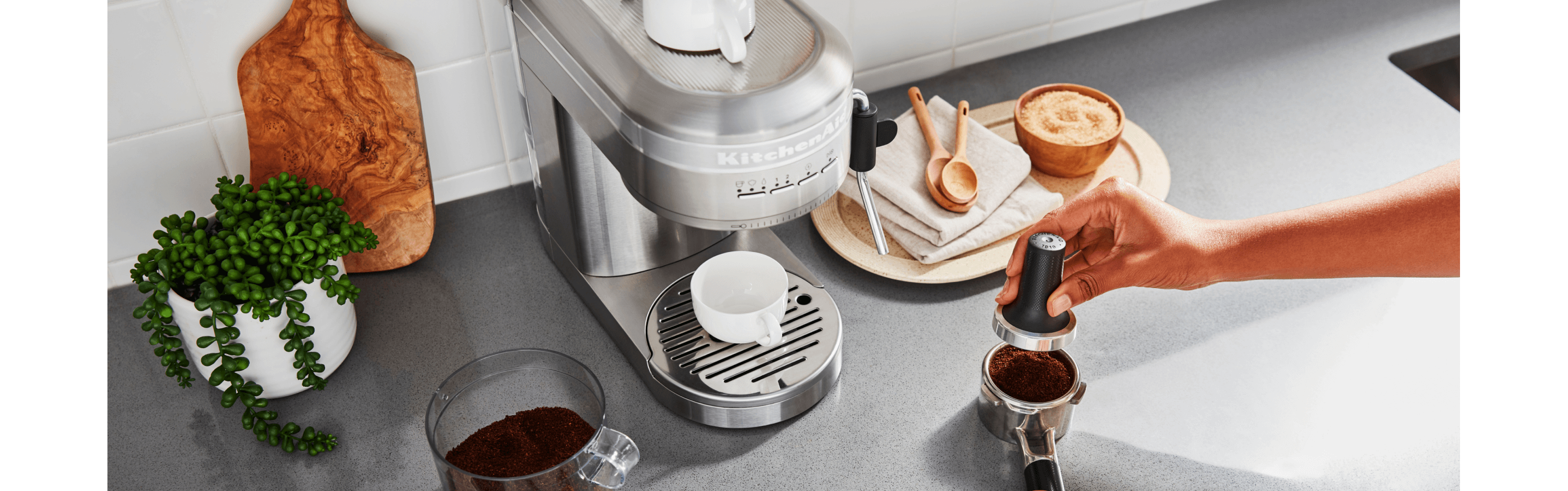 An image of a KitchenAid® Espresso machine, with a person pressing grounds into it.
