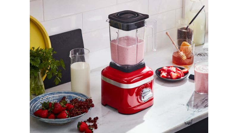Red K150 Blender with a fresh smoothie, beside a plate of cut strawberries and jar of honey.