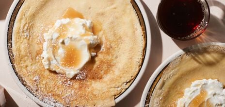 Plates of perfectly toasted crepes sit, dusted with powdered sugar. Each has a big dollop of melting butter in the center while maple syrup sits ready on the side.
