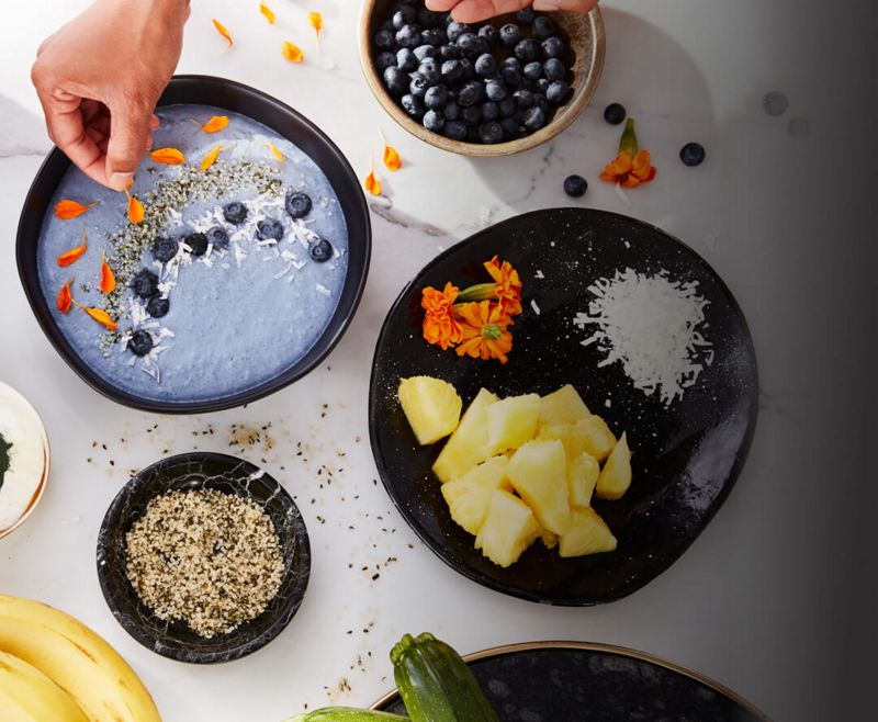 Overhead shot of a delicious-looking bowl of blue blueberry yogurt being topped with accompaniments.