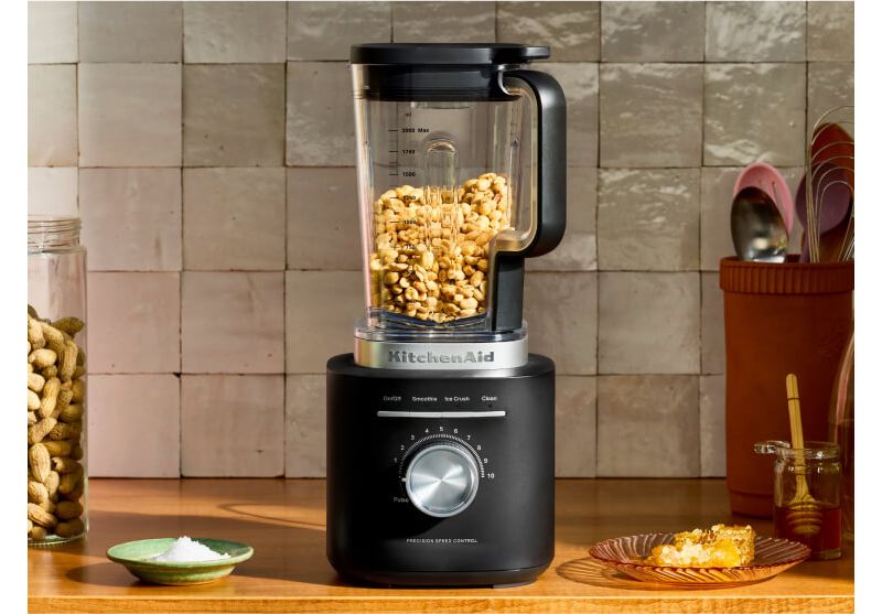 A black KitchenAid® Pure Power Blender with whole peanuts inside and a tray of sea salt and homemade peanut butter next to it 