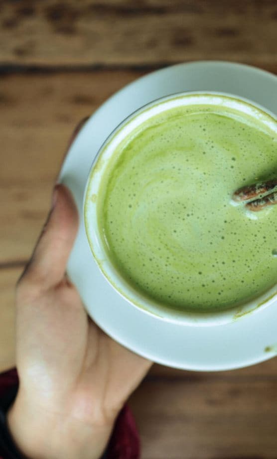 Soft hands holding a teacup filled with matcha latte and a cinnamon stick.