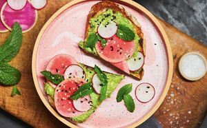 A pink plate with avocado toast topped with watermelon radishes and garnished with sesame and mint.