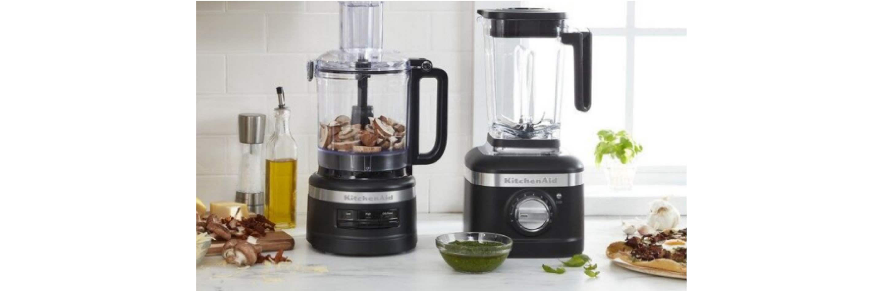 A KitchenAid food processor and blender on a countertop with different ingredients scattered around the counter. 