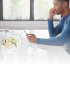 A young man looks at his phone in the background, in the foreground we see a clear pitcher with fruit and water