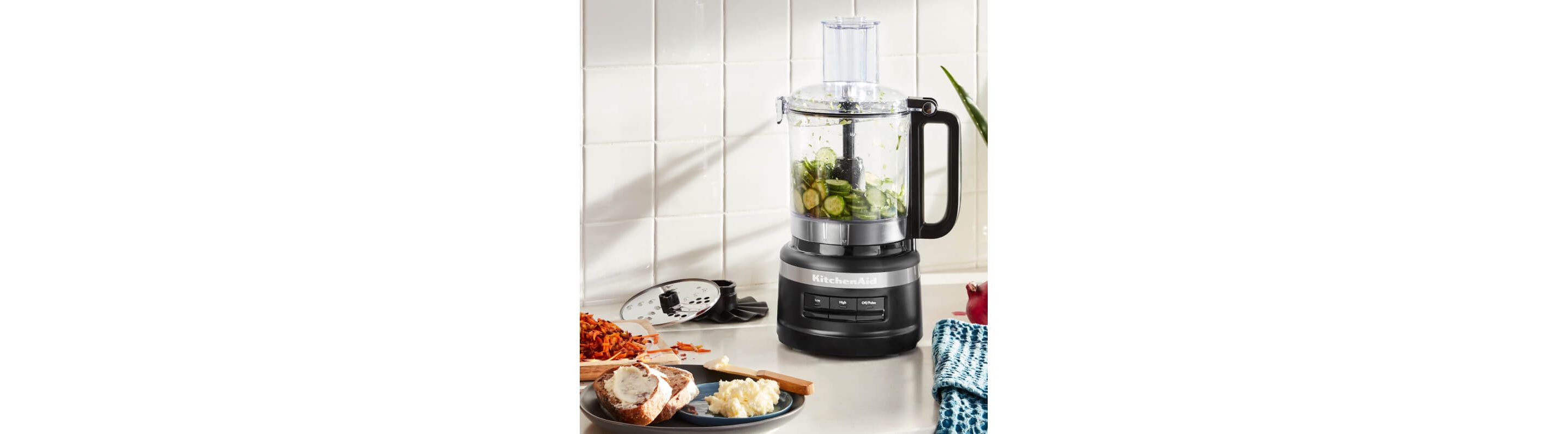 A Matte Black KitchenAid® food processor holding sliced cucumbers on a white countertop.