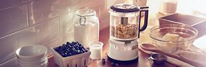 A White KitchenAid® food processor on a wooden countertop with blueberries and other ingredients.