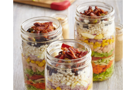 Three tall, glass mason jars filled with chopped ingredients for to-go salad.