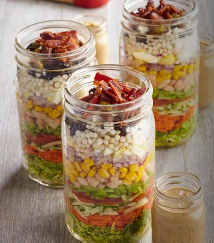Three tall, glass mason jars filled with chopped ingredients for to-go salad.