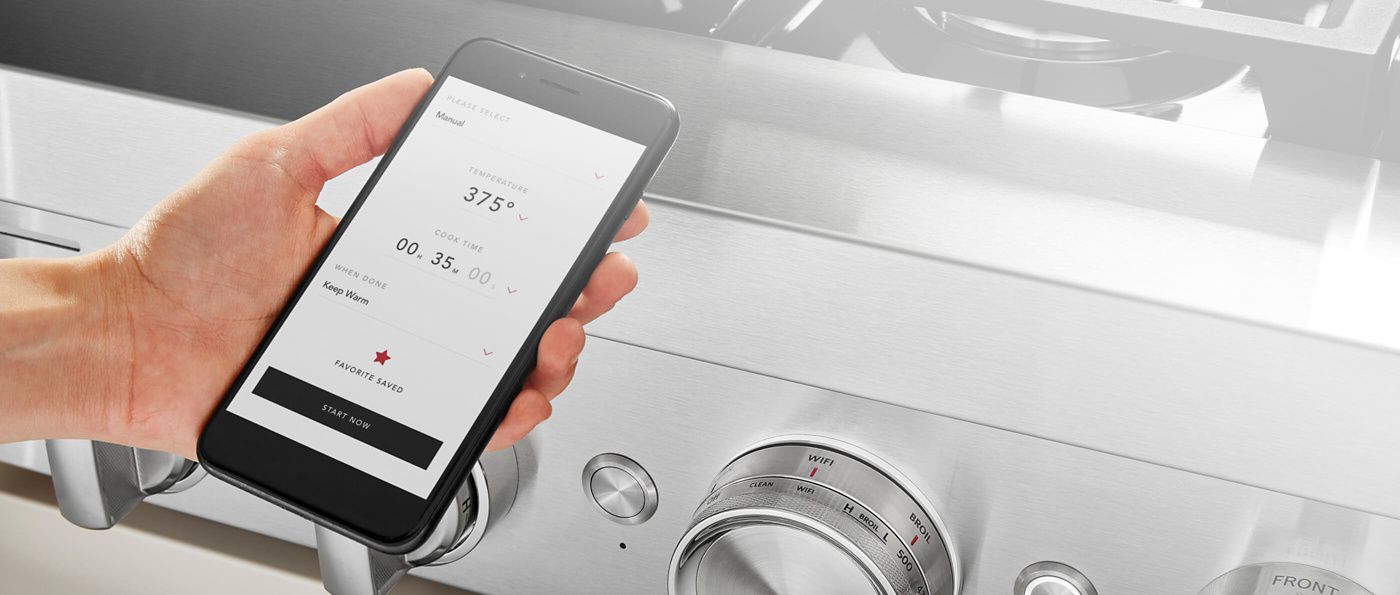 Person holding a smartphone in front of a range turning the temperature dial.