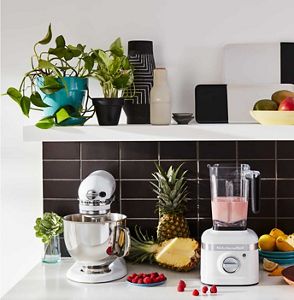 A white KitchenAid stand mixer next to a white KitchenAid blender, surrounded by fruits.