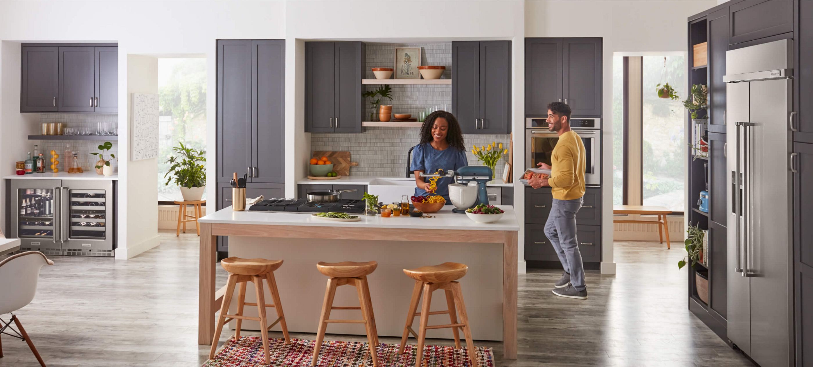 Two people in a large, modern kitchen, cooking different ingredients on KitchenAid® appliances.