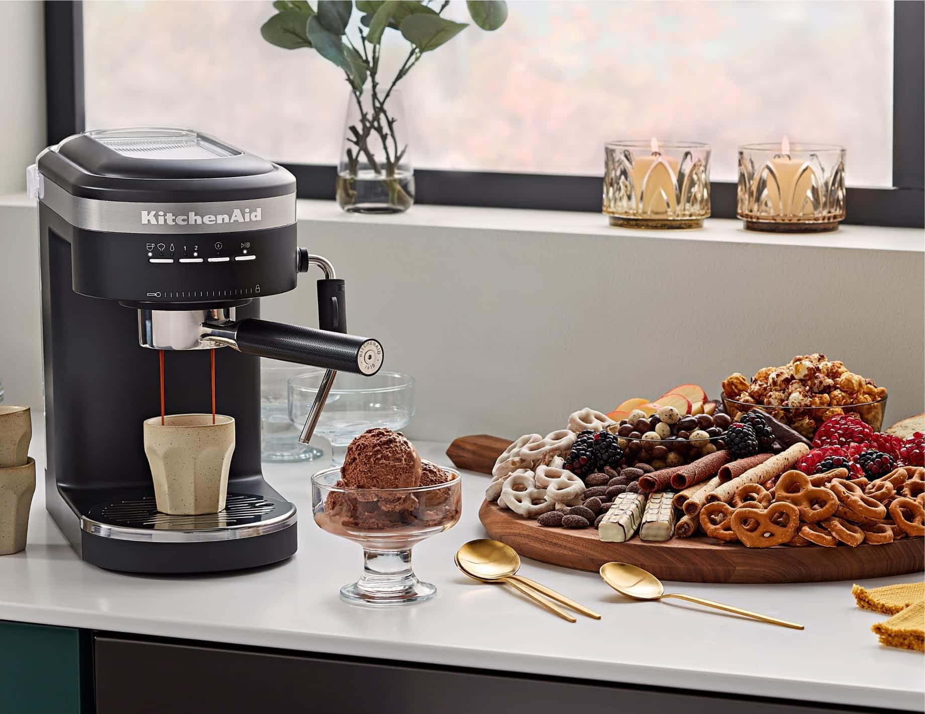 A KitchenAid® espresso machine brewing a cup of espresso next to a dessert charcuterie and cup of ice cream.