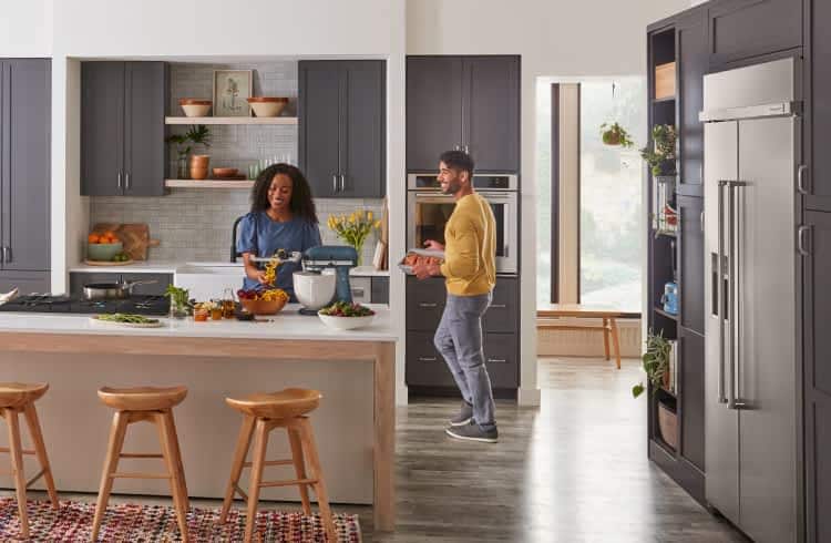 Two people in a large, modern kitchen, cooking different ingredients on KitchenAid® appliances.