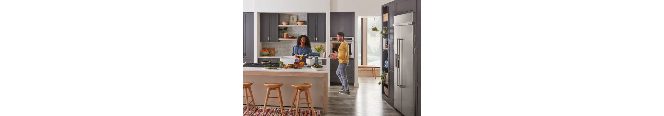Two people in a large, modern kitchen, cooking different ingredients on KitchenAid® appliances.