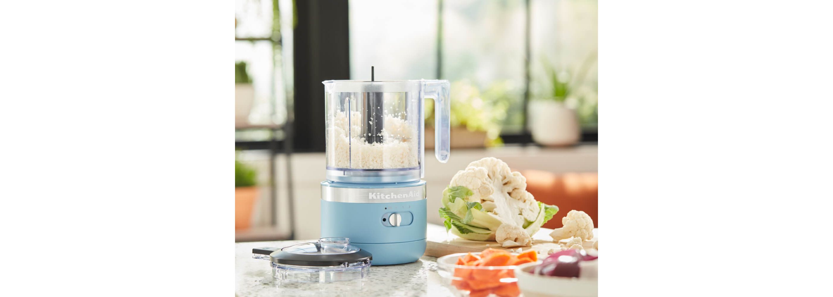 Chopping cauliflower florets in KitchenAid® 3.5 Cup Food Chopper in Blue Velvet, cauliflower and carrots on countertop