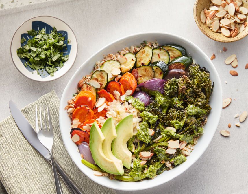 White bowl containng fresh avocado, carrots, cauliflower, zucchini, herbs, nuts and grains.