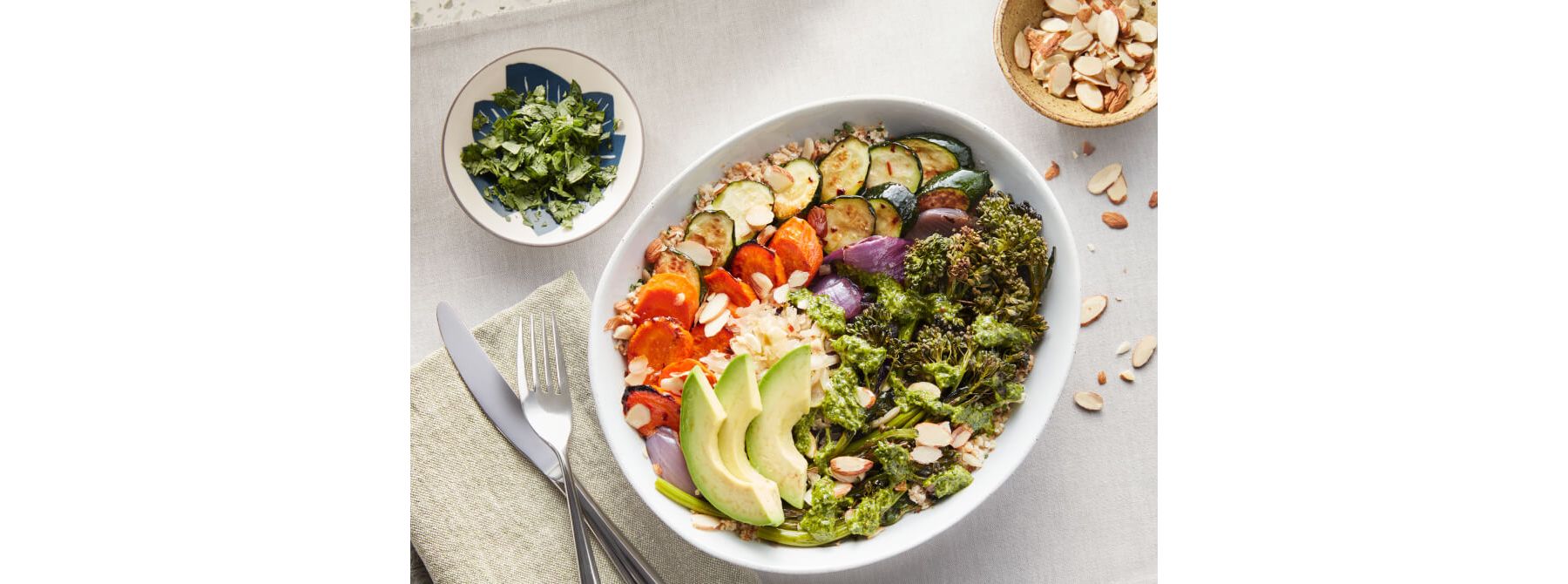 White bowl containng fresh avocado, carrots, cauliflower, zucchini, herbs, nuts and grains.