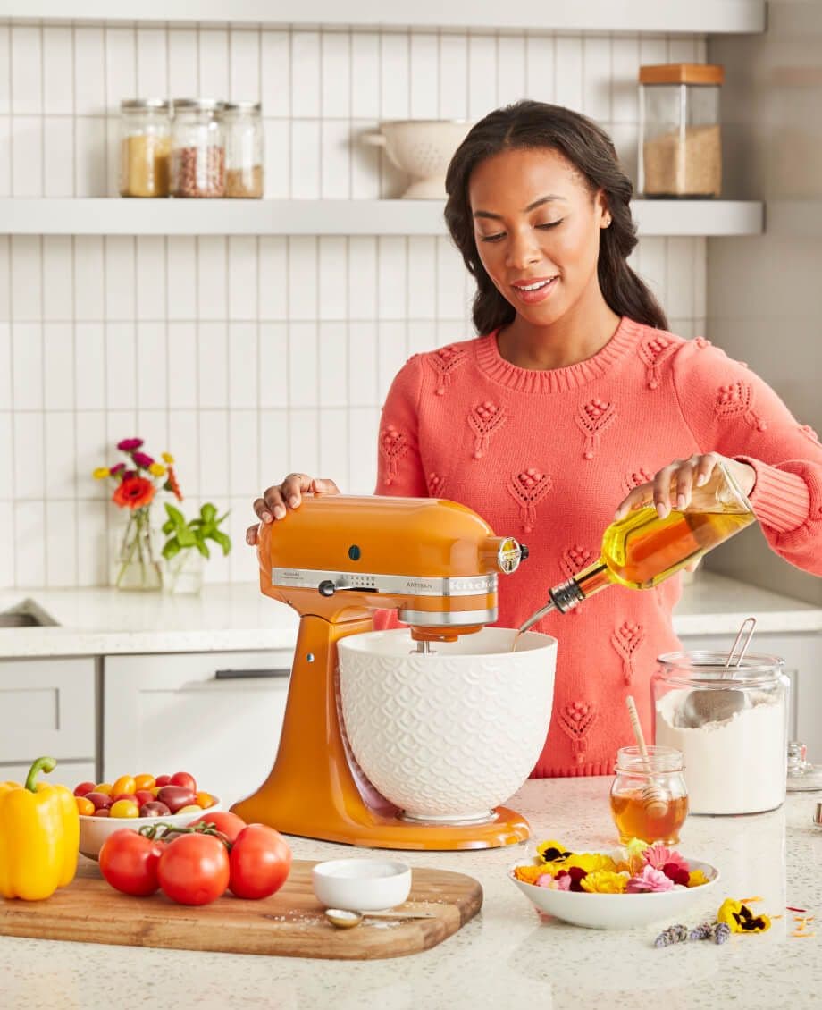 Woman in orange sweater pouring oil into white ceramic bowl of KitchenAid® Stand Mixer in Honey.