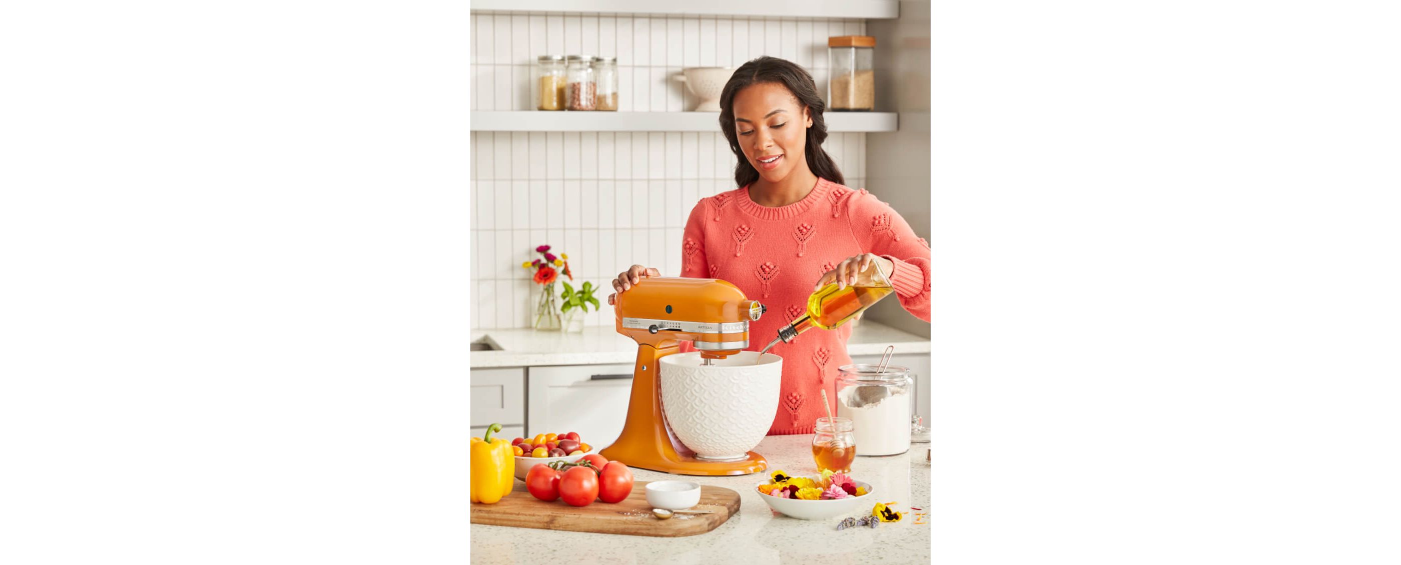 Woman in orange sweater pouring oil into white ceramic bowl of KitchenAid® Stand Mixer in Honey.