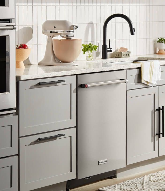 Kitchen setting with KitchenAid® Stand Mixer sitting on white countertop and stainless color KitchenAid® Dishwasher set in white cabinetry.
