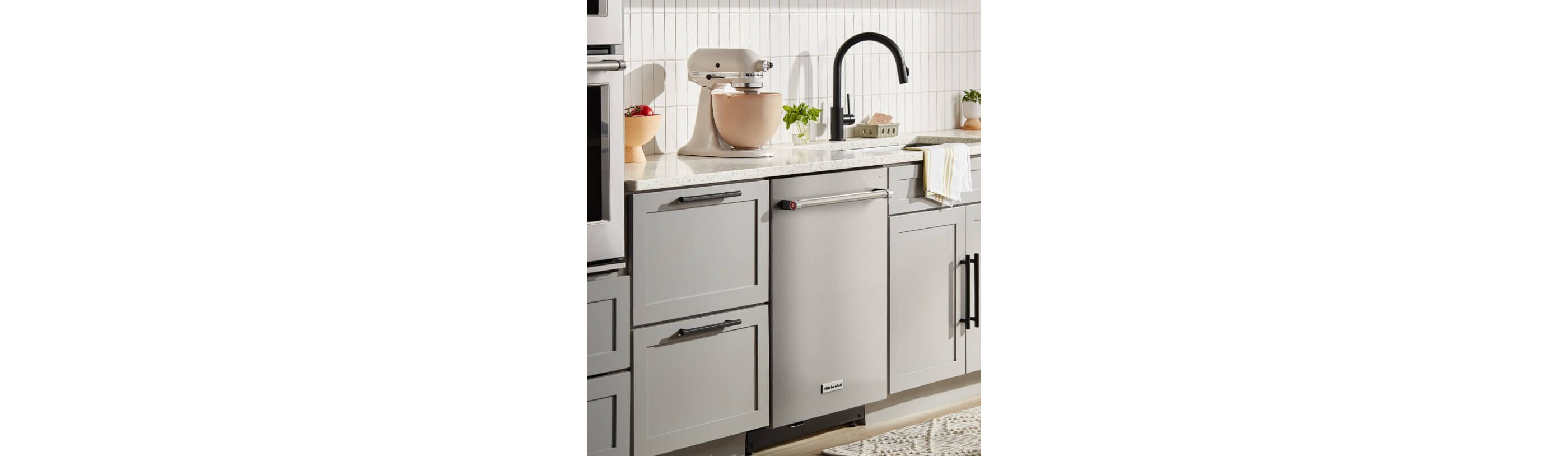 Kitchen setting with KitchenAid® Stand Mixer sitting on white countertop and stainless color KitchenAid® Dishwasher set in white cabinetry.