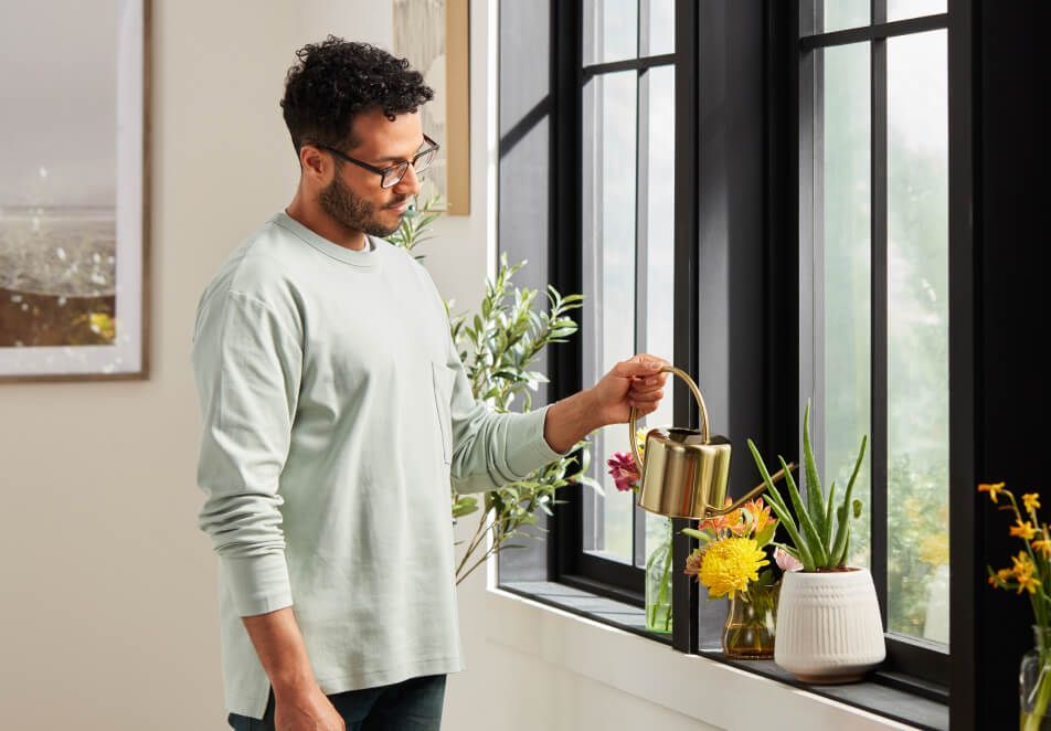 Man in grey t-shirt watering plants in windowsill.