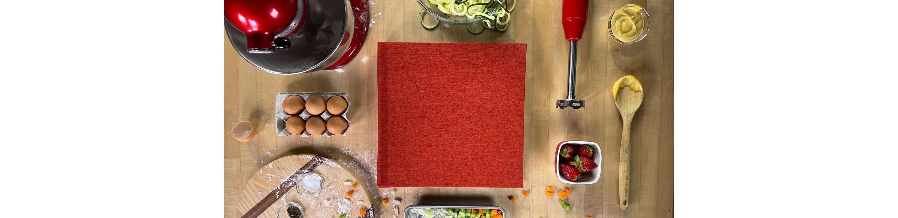 A kitchen countertop scattered with different ingredients and KitchenAid appliances with a red cookbook.