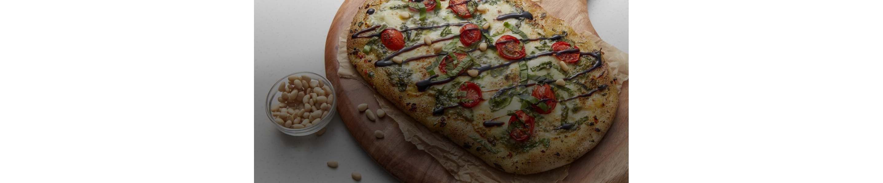A flatbread pizza on a wood serving board next to a bowl of pine nuts. 