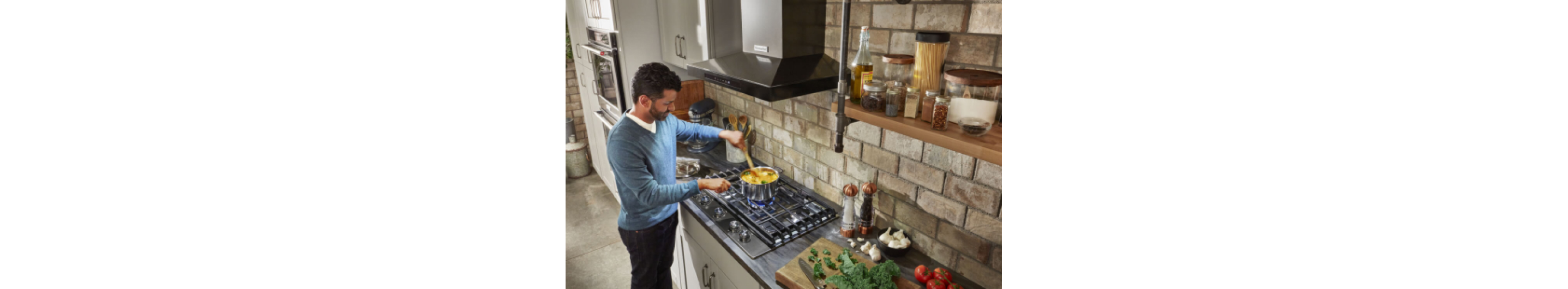 A person stirring food in a pot on a cooktop with a KitchenAid® Vent Hood above.