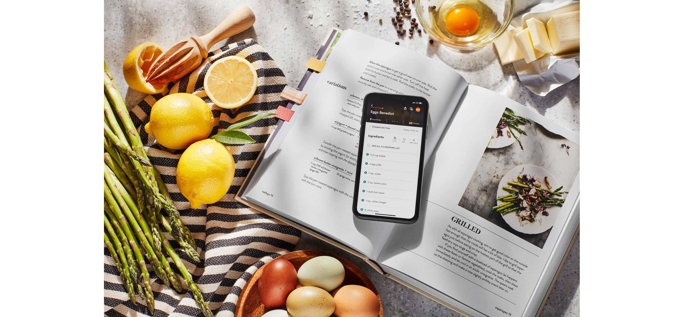 A phone opened up to an eggs benedict recipe resting on an open book&#x27;s grilled asparagus recipe