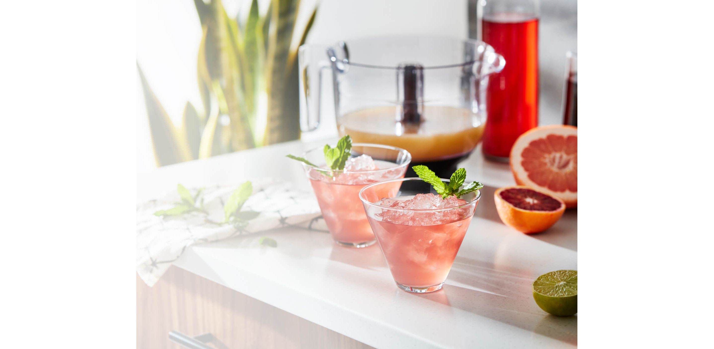 Two freshly made grapefruit cocktails resting on a bright kitchen counter with assorted citruses.