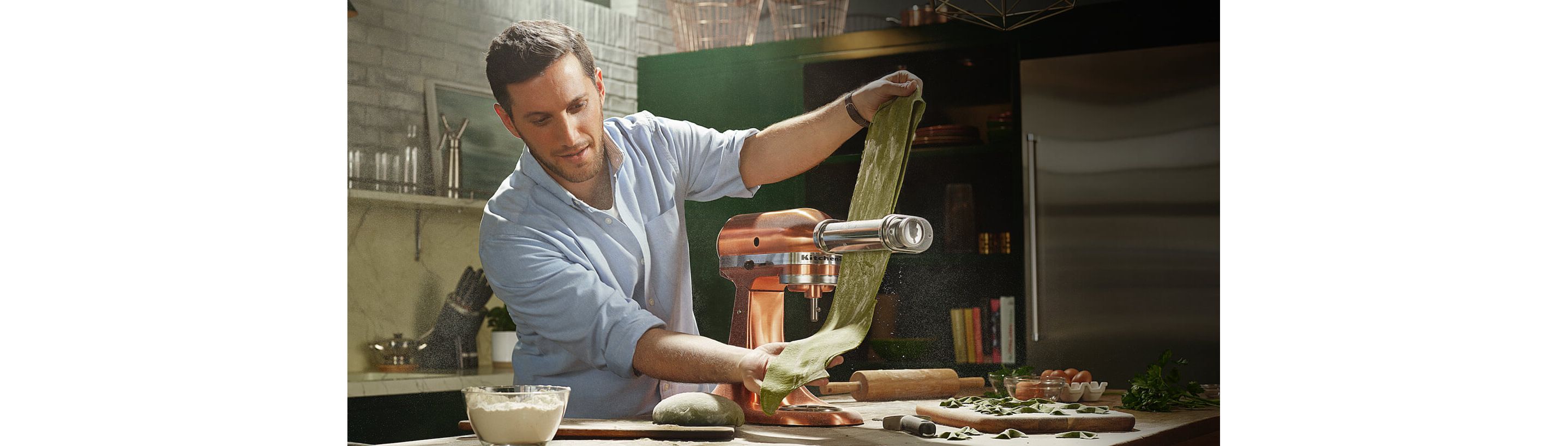 A man running green pasta dough through a pasta sheet roller attached to a Copper Stand Mixer