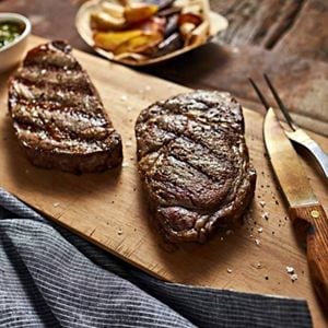 Two grilled ribeye steaks resting on a wooden cutting board.