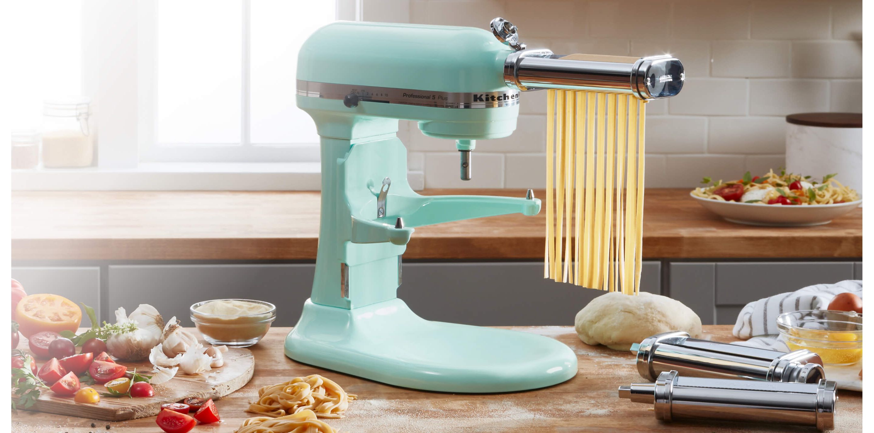 Aqua-colored Stand Mixer with pasta attachment, cutting pasta sheets into linguine on a counter adorned with fresh vegetables.