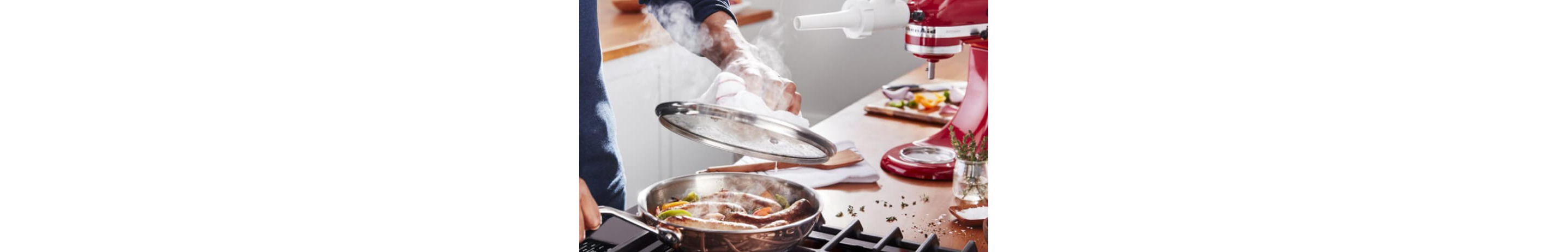A man cooking juicy bratwursts with sautéed vegetables in a stainless steel pan over a stove.