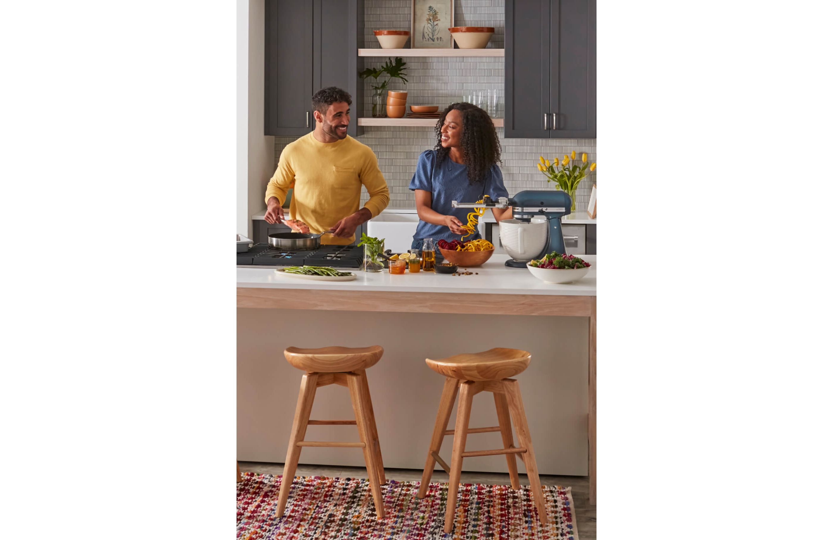 A man and woman cooking, featuring a stand mixer and gas cooktop.