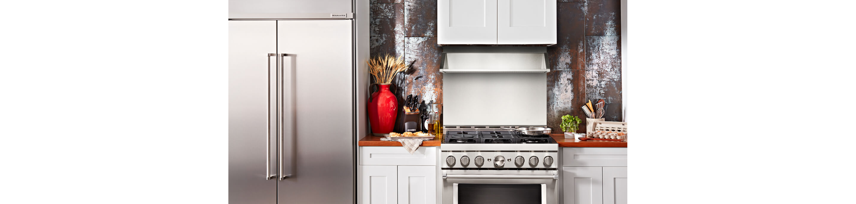 Cropped picture of a kitchen section, with stainless steel refrigerator, commercial-style range and backguard, as well as various cooking items. 