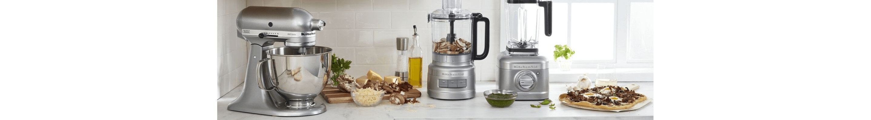 A KitchenAid® Stand Mixer, Food Processor and Blender on a white countertop, surrounded by different ingredients on a cutting board and a prepared breakfast pizza.