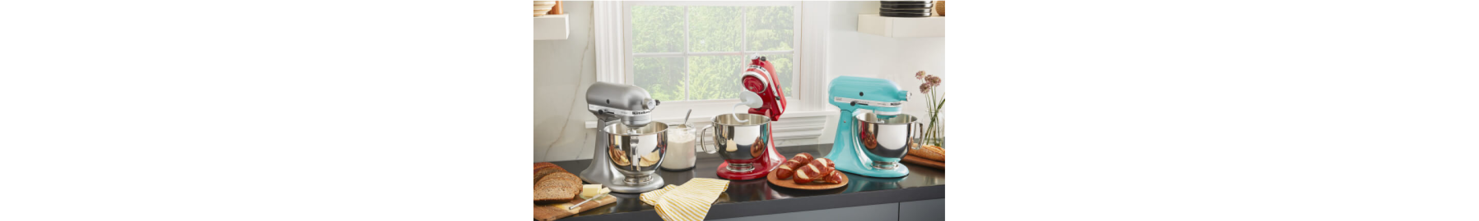 A trio of KitchenAid® Stand Mixers on a countertop with fresh baked bread on plates. 