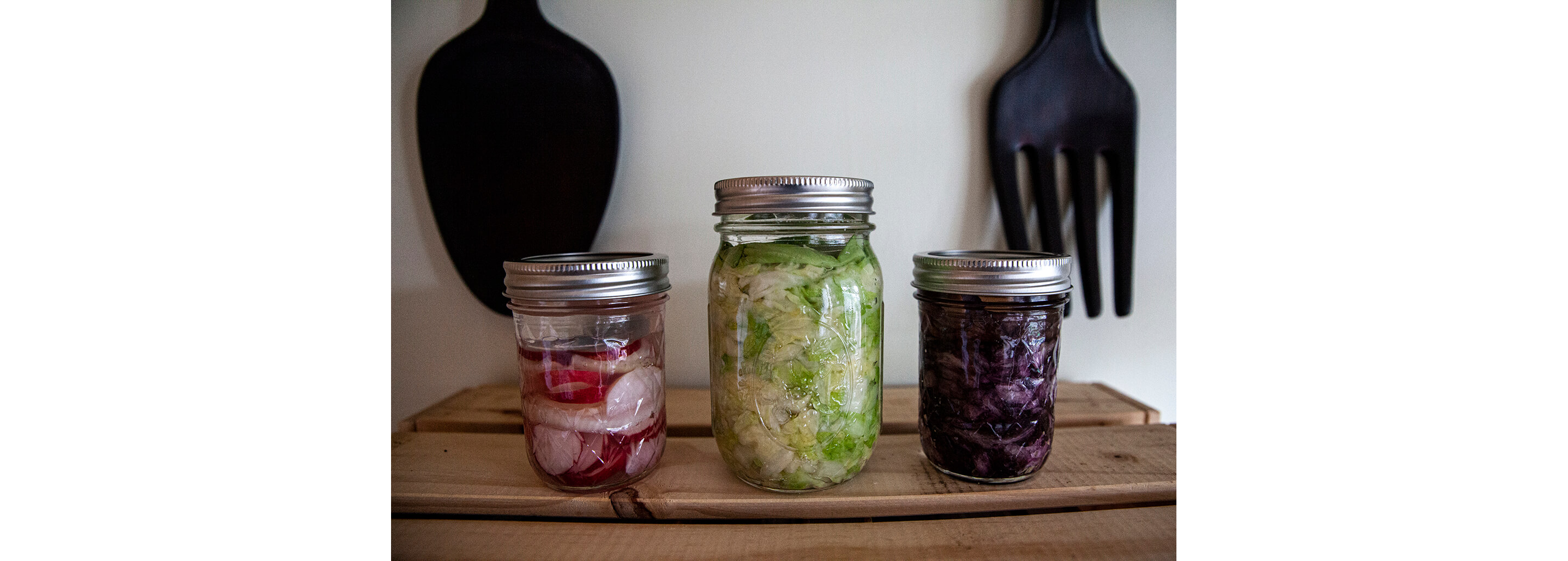 Three glass jars holding fermented radishes, red cabbage and green cabbage.