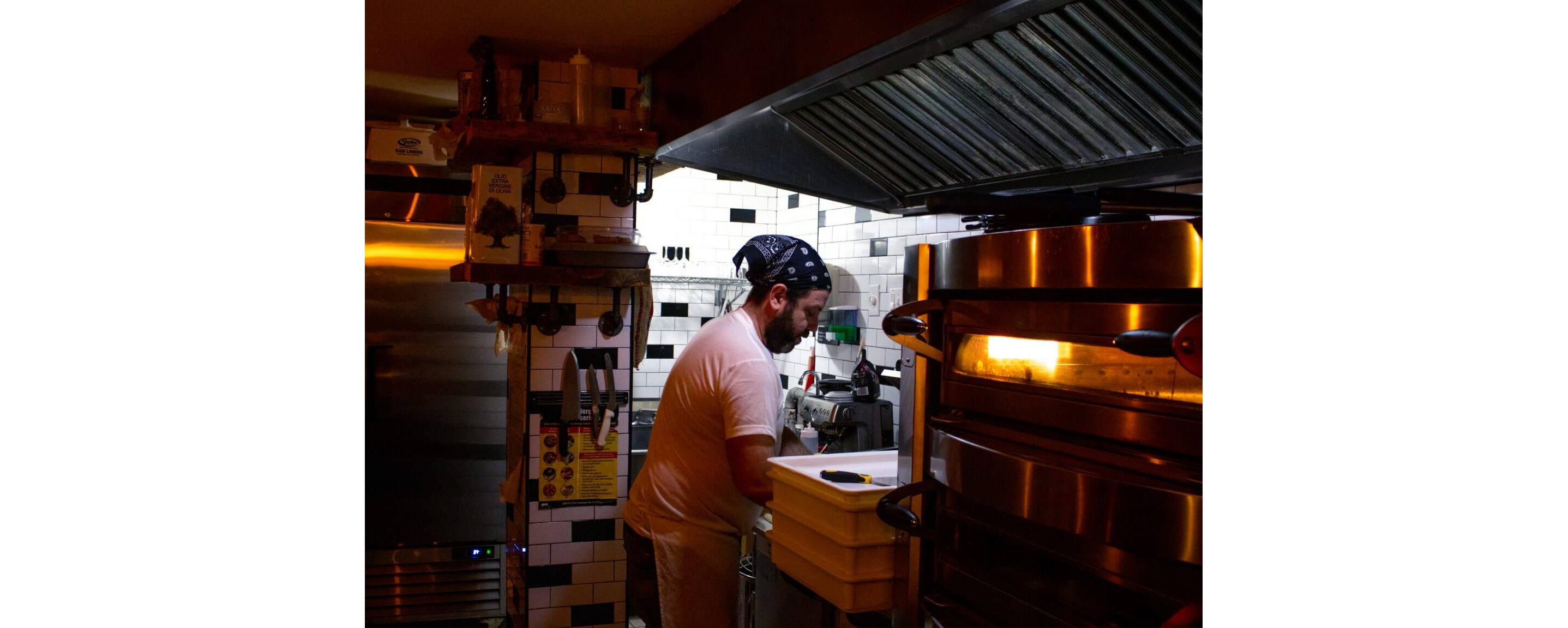 A man standing in his restaurant's kitchen working diligently over a pizza.