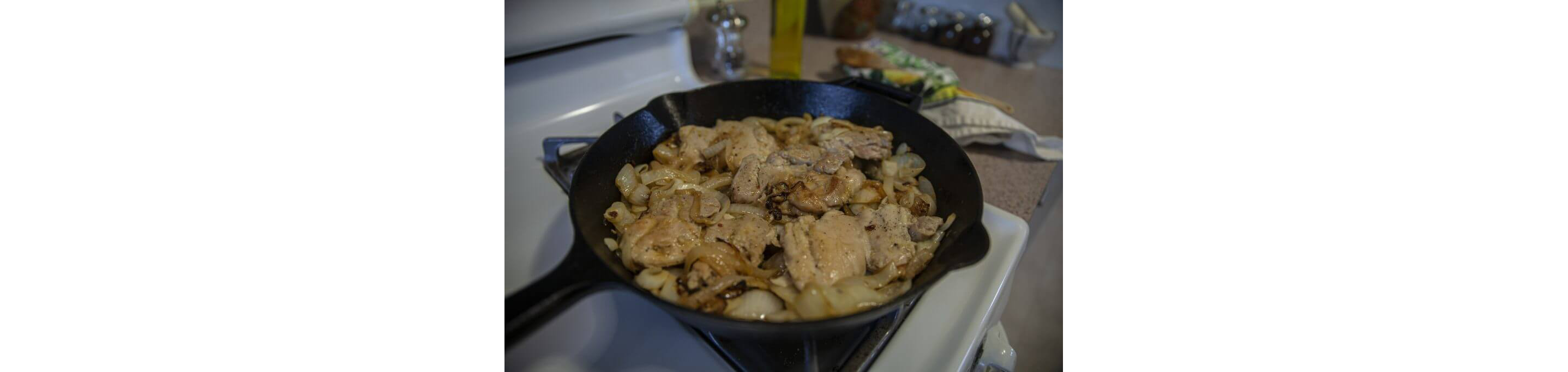 A stove with a cast iron skillet holding chicken thighs and caramelized onions.