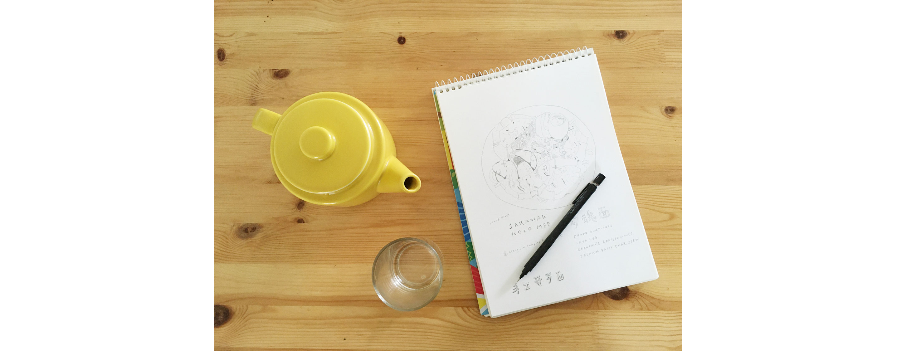A sketch book resting on a table with a pot of tea.
