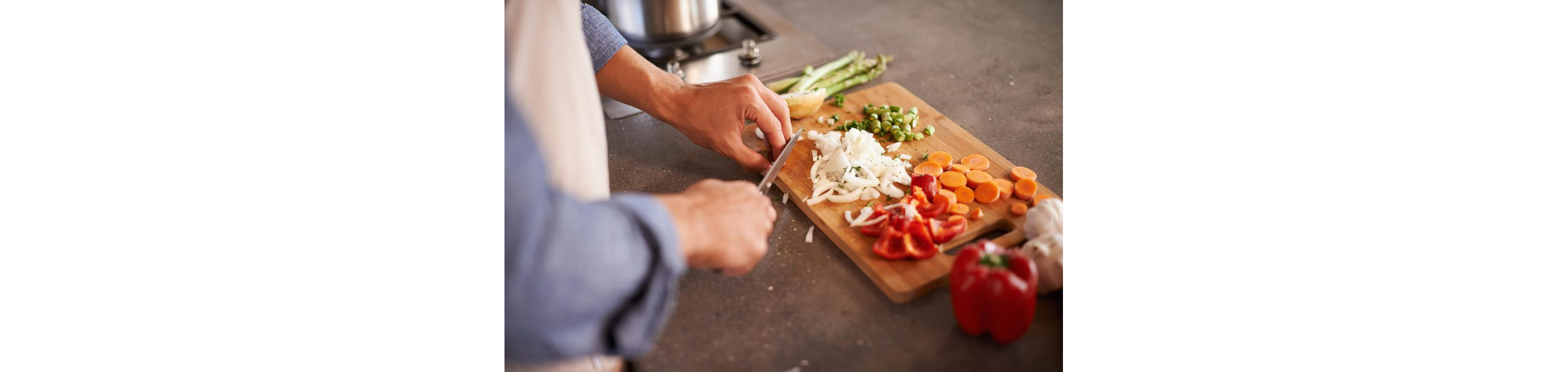 A person dicing white onions on a wooden cutting board along with diced asparagus and tomatoes.