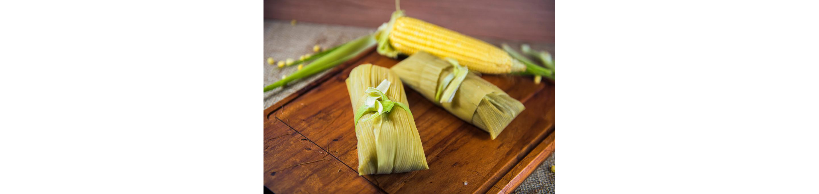 Pamonha resting on a wooden cutting board next to a peeled corn on the cob.