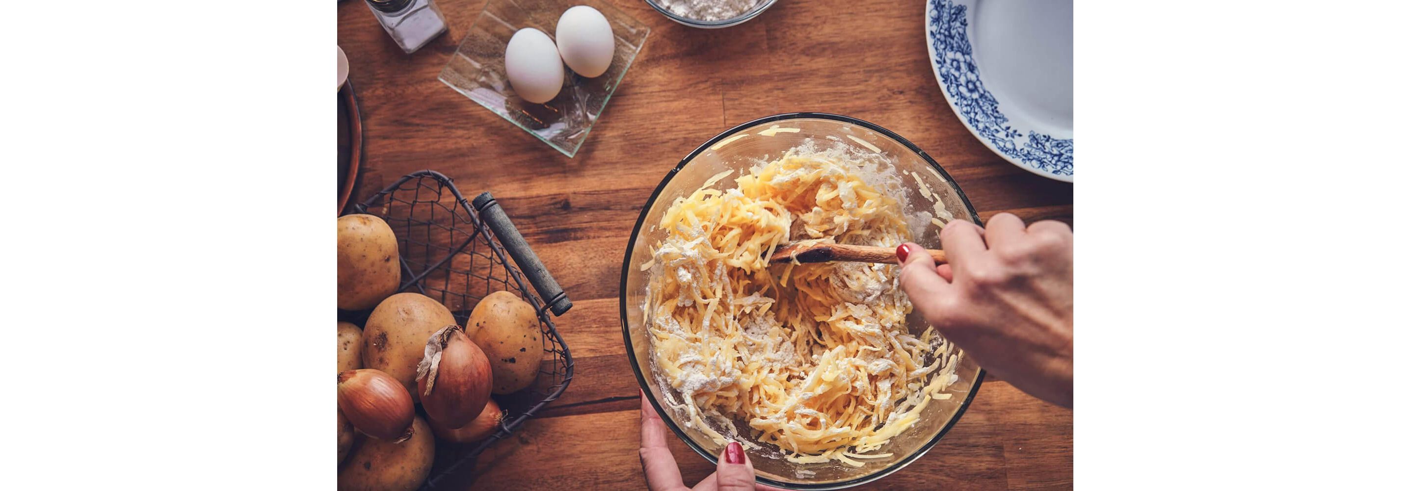 Strong hands stirring shredded potatoes in a glass bowl with a wooden spoon.