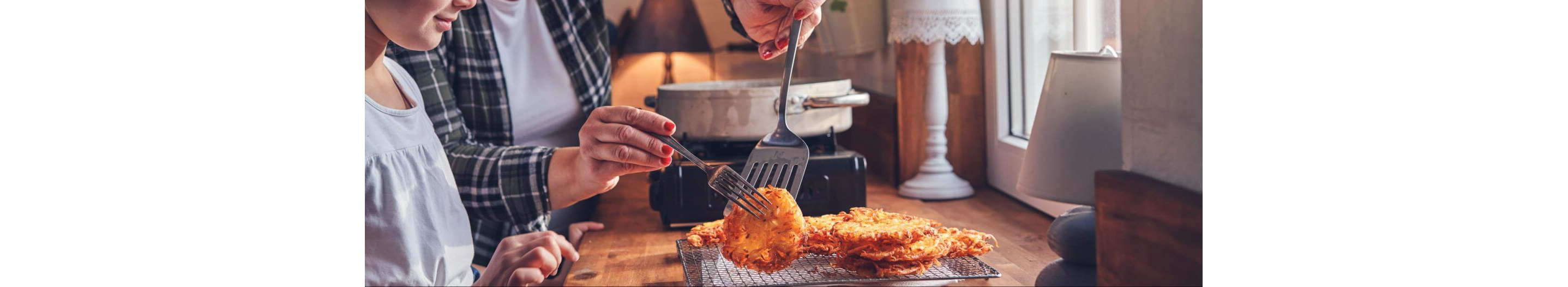 A young girl watching her mother gently move potato pancakes to a cooling rack.