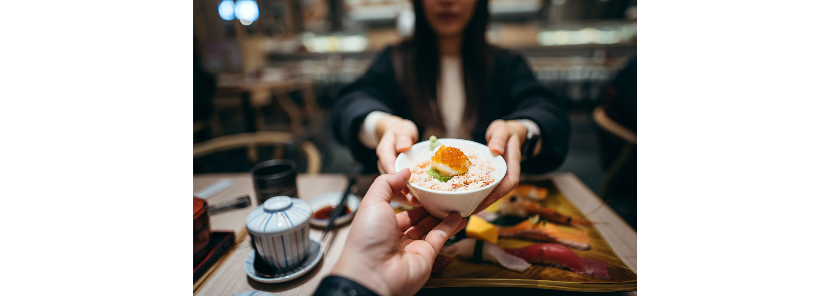 A person handing a small bowl filled with seafood donburi to a young woman.